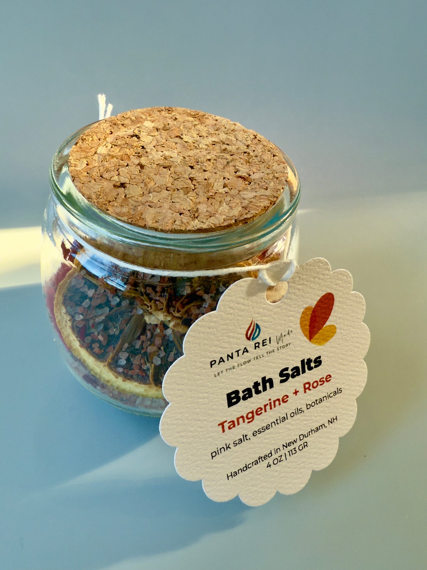 Glass jar with cork lid filled with pink salt, dried citrus slices, and rose petals, with product tag attached.