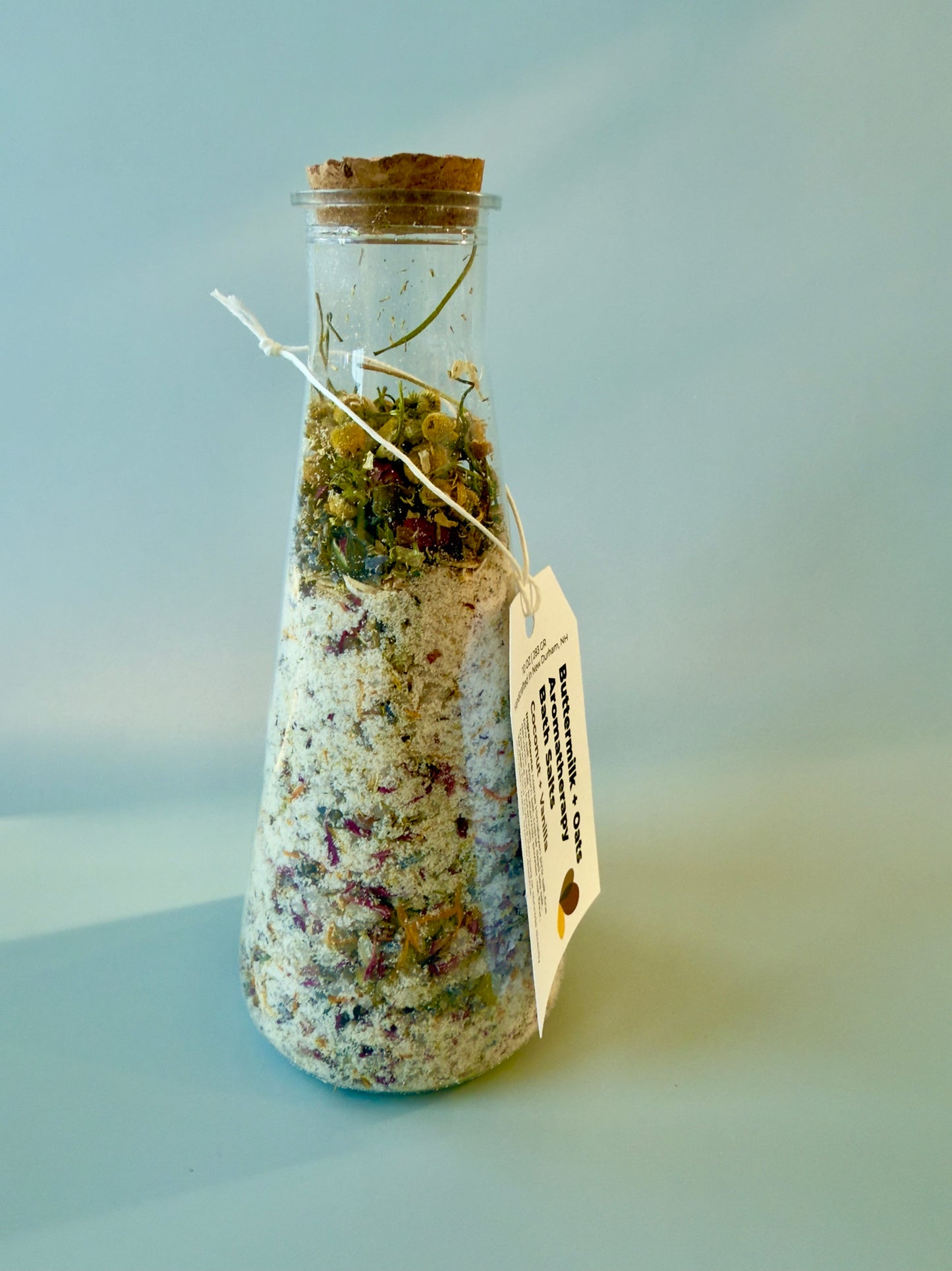 Tall conical glass bottle with cork top filled with coconut-vanilla bath salts and layered botanicals.