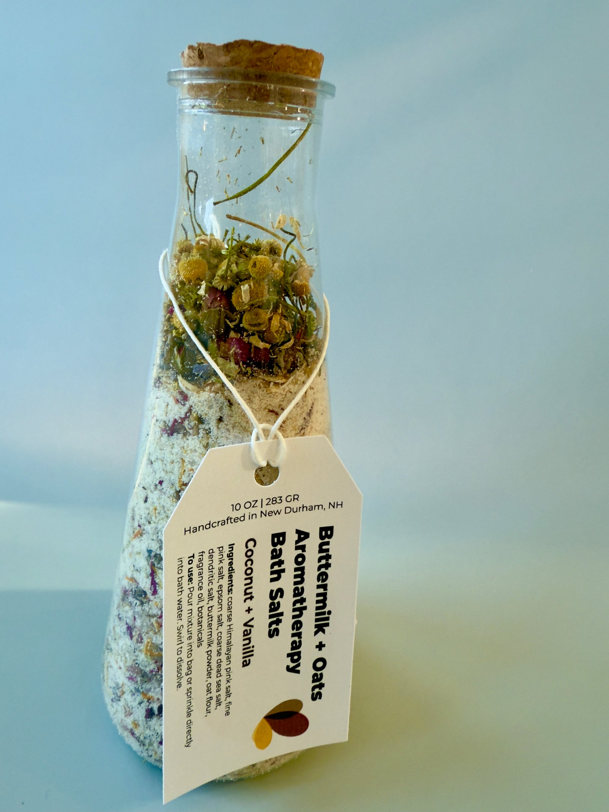 Close-up of Coconut + Vanilla bath salts bottle with chamomile botanicals and product tag.