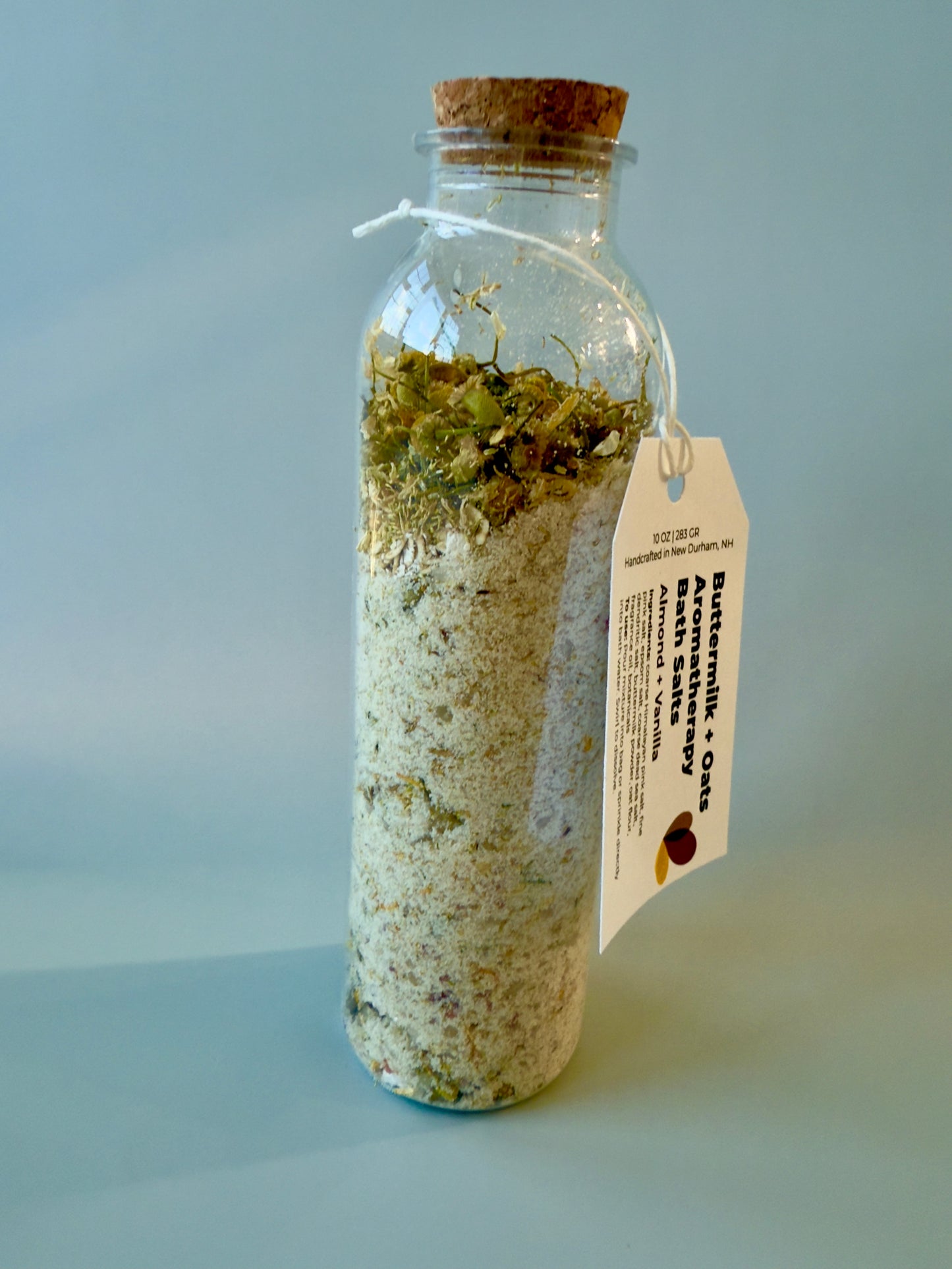Tall round glass bottle with cork top filled with buttermilk and oat bath salts and chamomile botanicals.