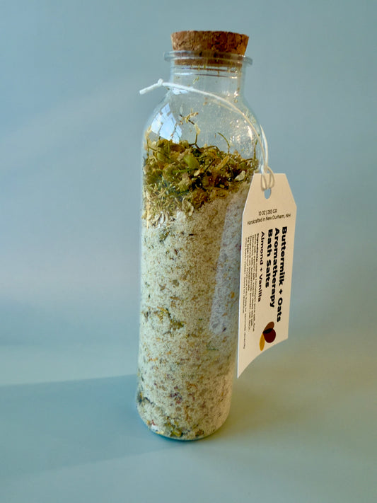 Tall round glass bottle with cork top filled with buttermilk and oat bath salts and chamomile botanicals.