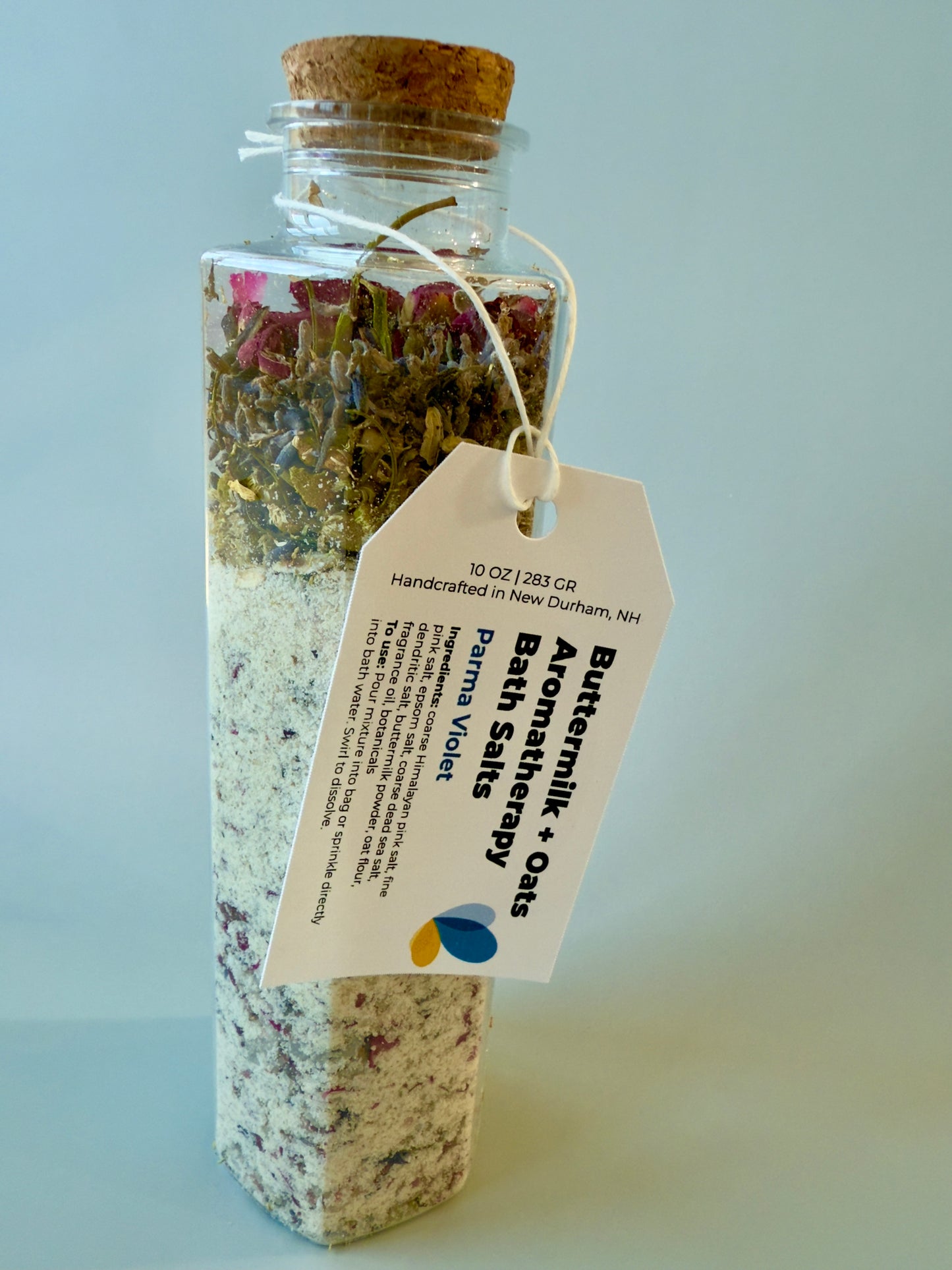 Tall square glass bottle with cork top, filled with buttermilk and oat bath salts and layered florals.