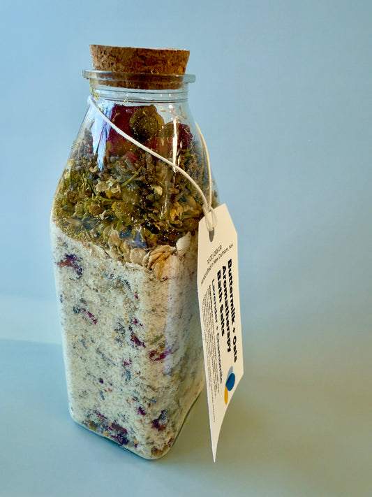 Square glass bottle with cork top filled with layered buttermilk and oat bath salts and visible dried botanicals.