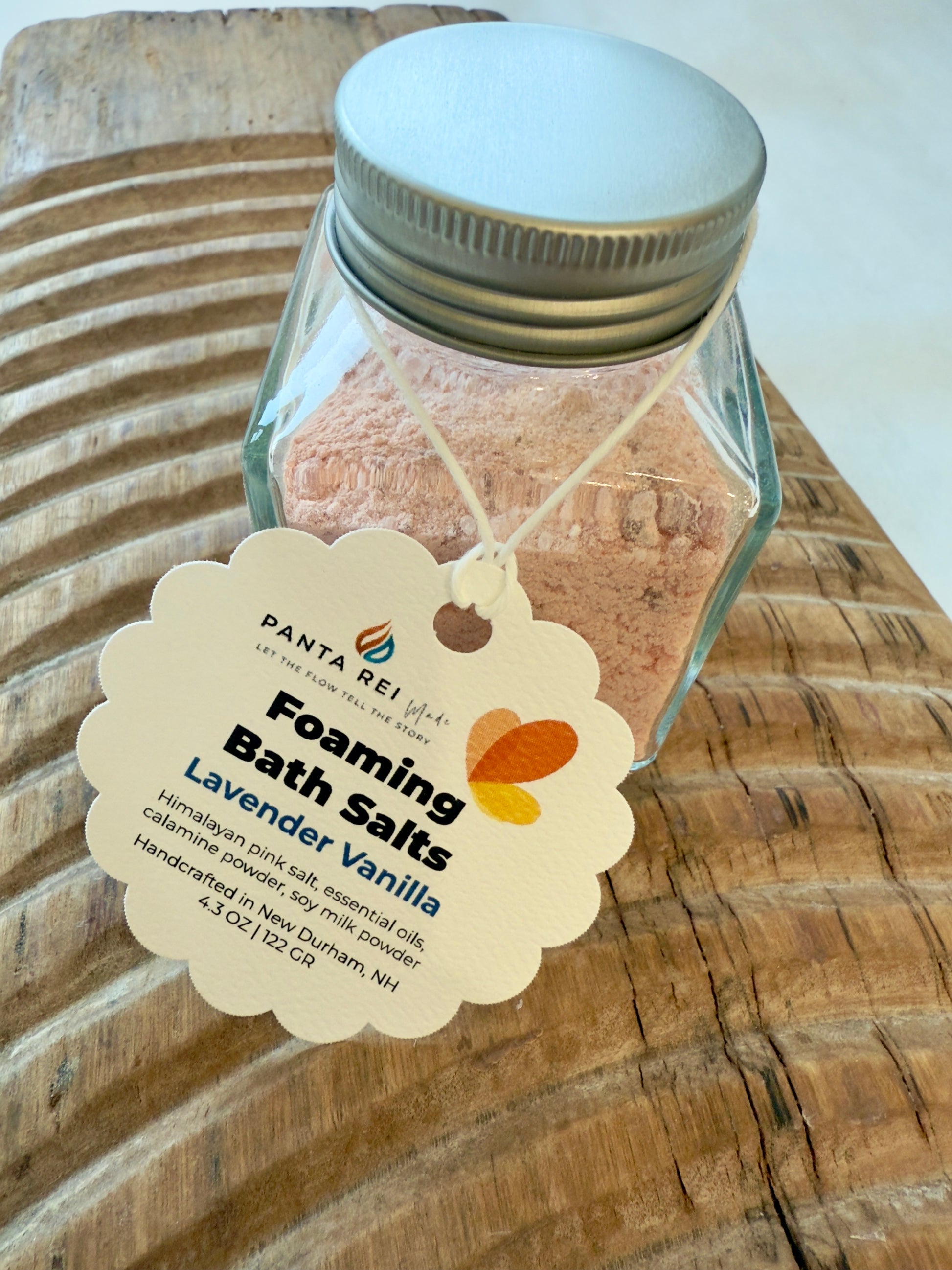 Square glass jar with metal lid filled with pink foaming bath salts and product tag.