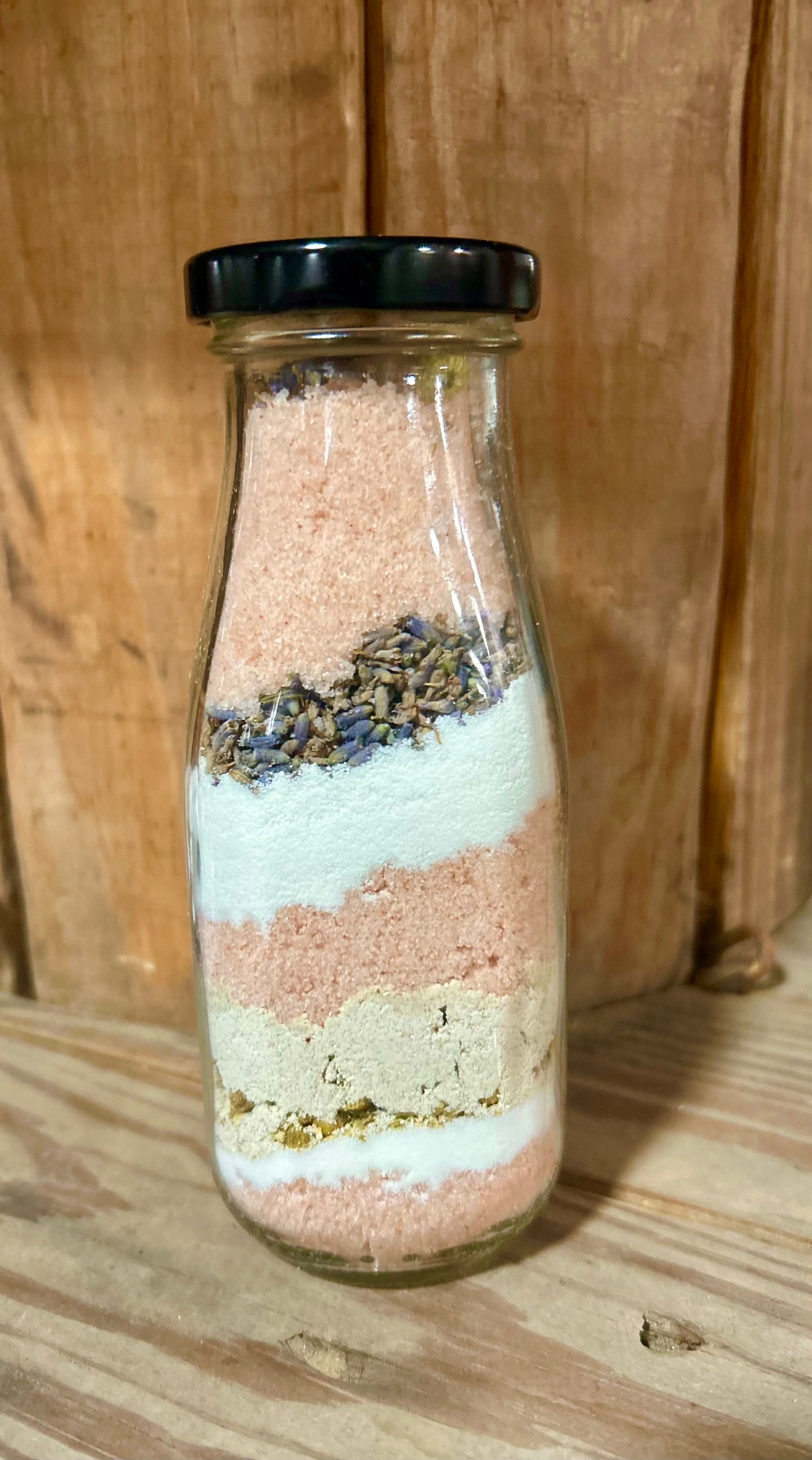 Layered milk and salt aromatherapy bath in a clear glass milk bottle with black cap, showing pink salt, coconut milk powder, oatmeal, and lavender buds.