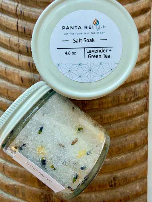 Glass jar of Lavender + Green Tea Salt Soak with botanicals on carved wood background and white lid.