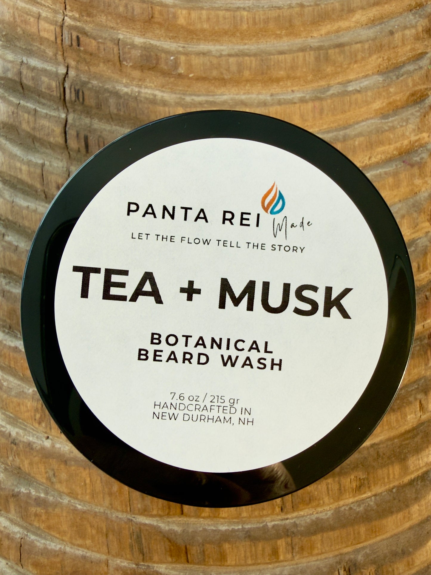 Tea + Musk Botanical Beard Wash jar with black lid on carved wood background.