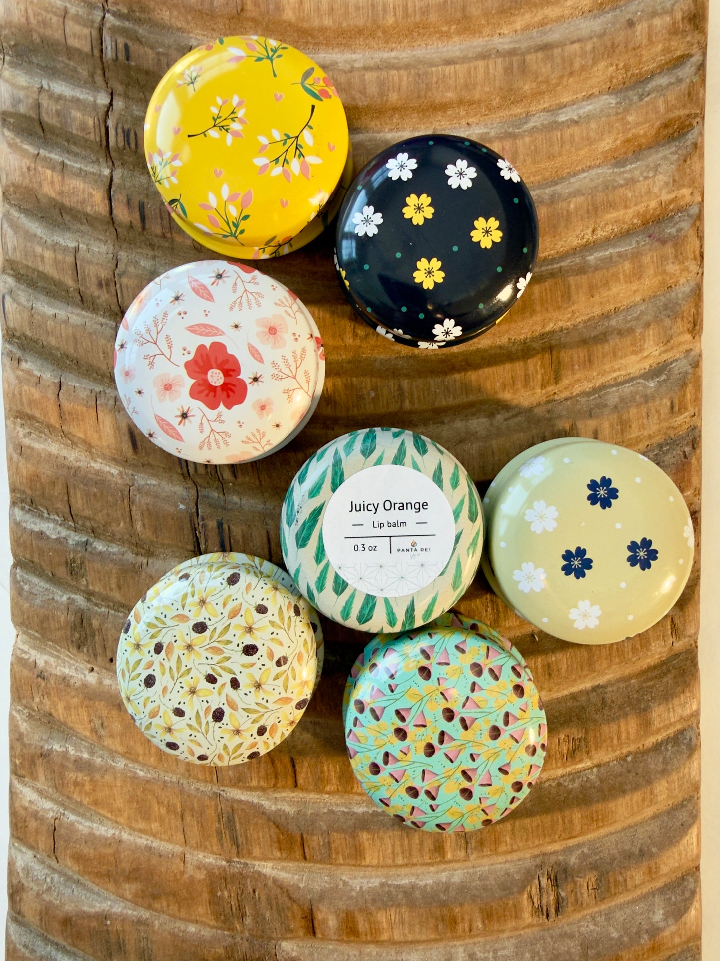 Floral tin of Juicy Orange lip balm with reusable metal packaging on textured wood surface.