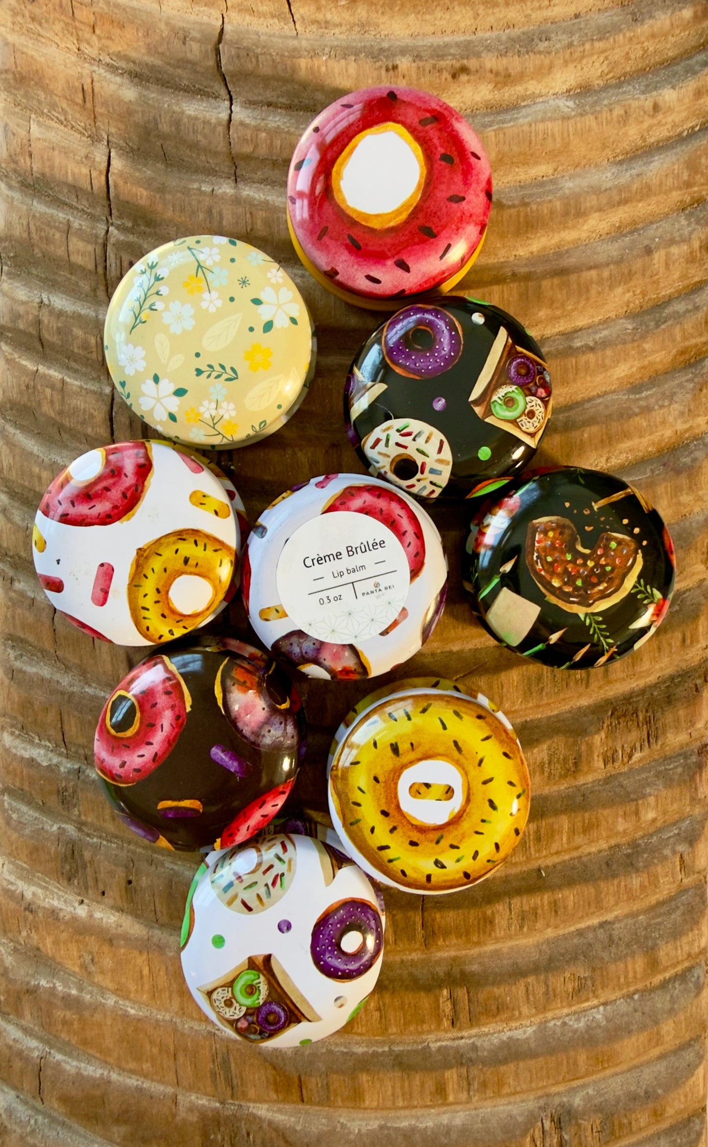 Crème Brûlée lip balm tin with donut-print design arranged among colorful patterned tins on carved wood surface.