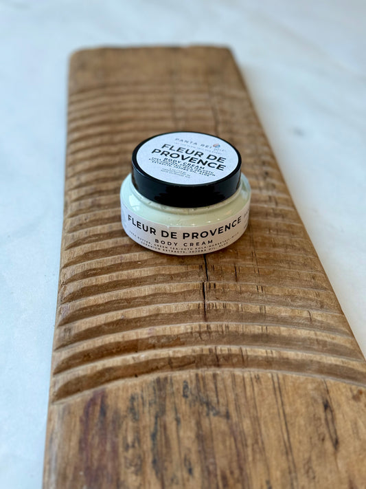 Glass jar of Fleur de Provence body cream with a black lid, filled with pale green cream and placed on a carved wooden board against a light background