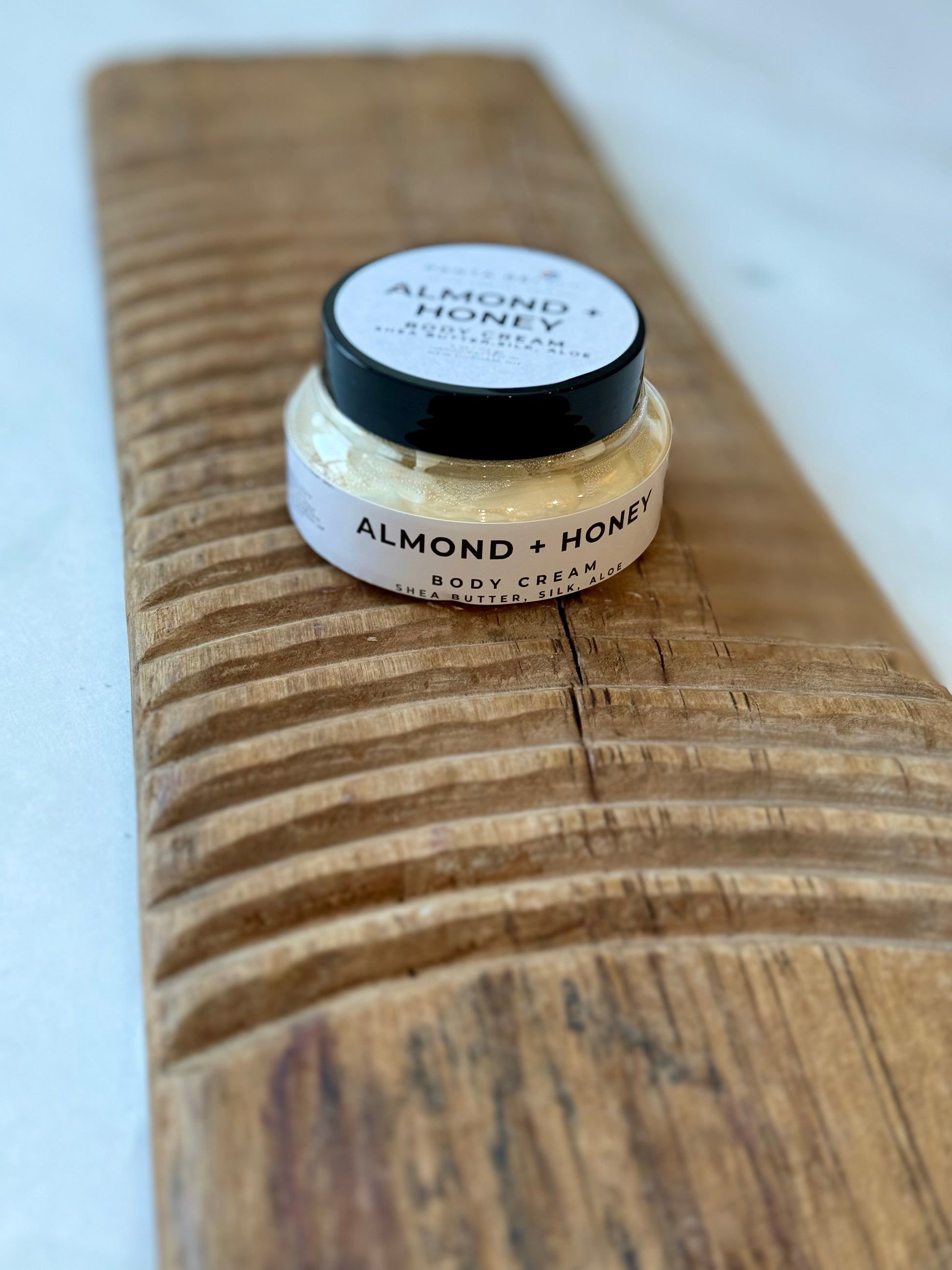 Glass jar of Almond + Honey body cream with a black lid, filled with pale cream and placed on a carved wooden board against a light background