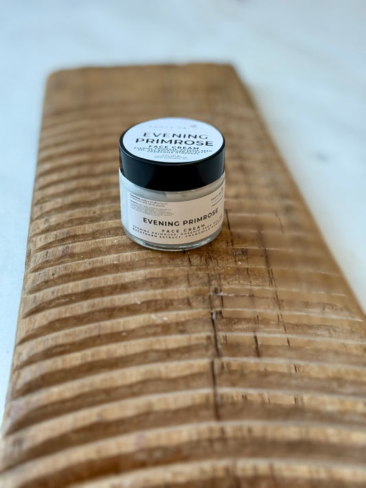 Small glass jar of Evening Primrose face cream with a black lid, placed on a carved wooden board against a light background