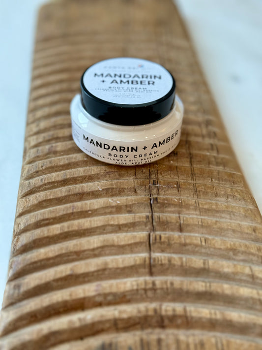 Jar of Mandarin + Amber body cream with a black lid, filled with white cream and placed on a carved wooden board against a light background