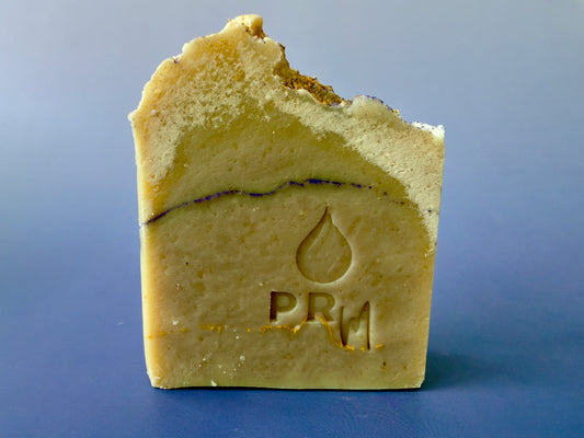 Lavender Cedarwood soap bar with PRM logo, gold and purple mica accents visible along the top, set against a blue background