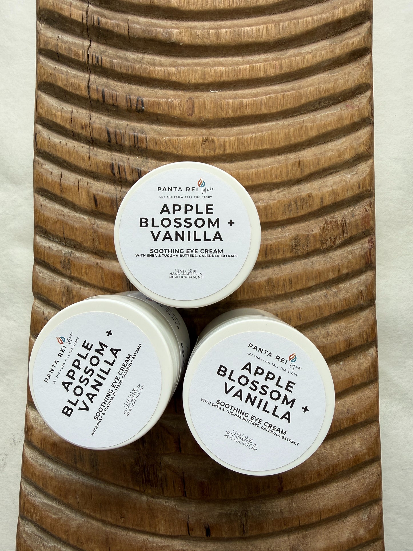 Three containers of Apple Blossom Vanilla eye cream on a wooden surface