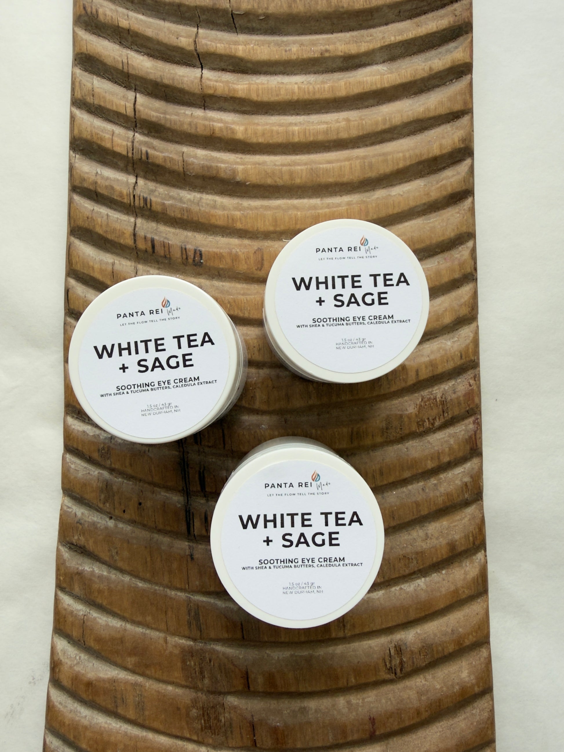 Three containers of 'White Tea + Sage' eye cream on a wooden board.
