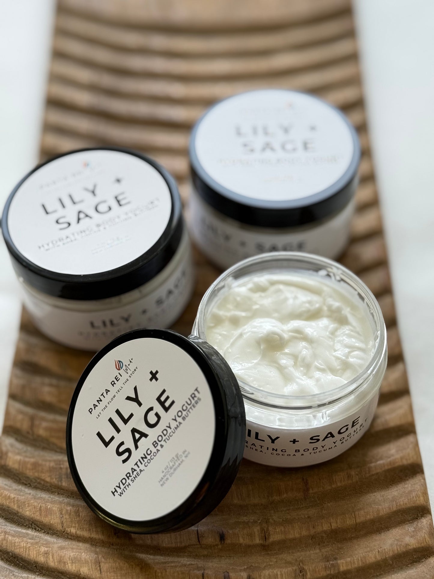 Lily + Sage hydrating body yogurt in white jars with black lids on a wooden tray.