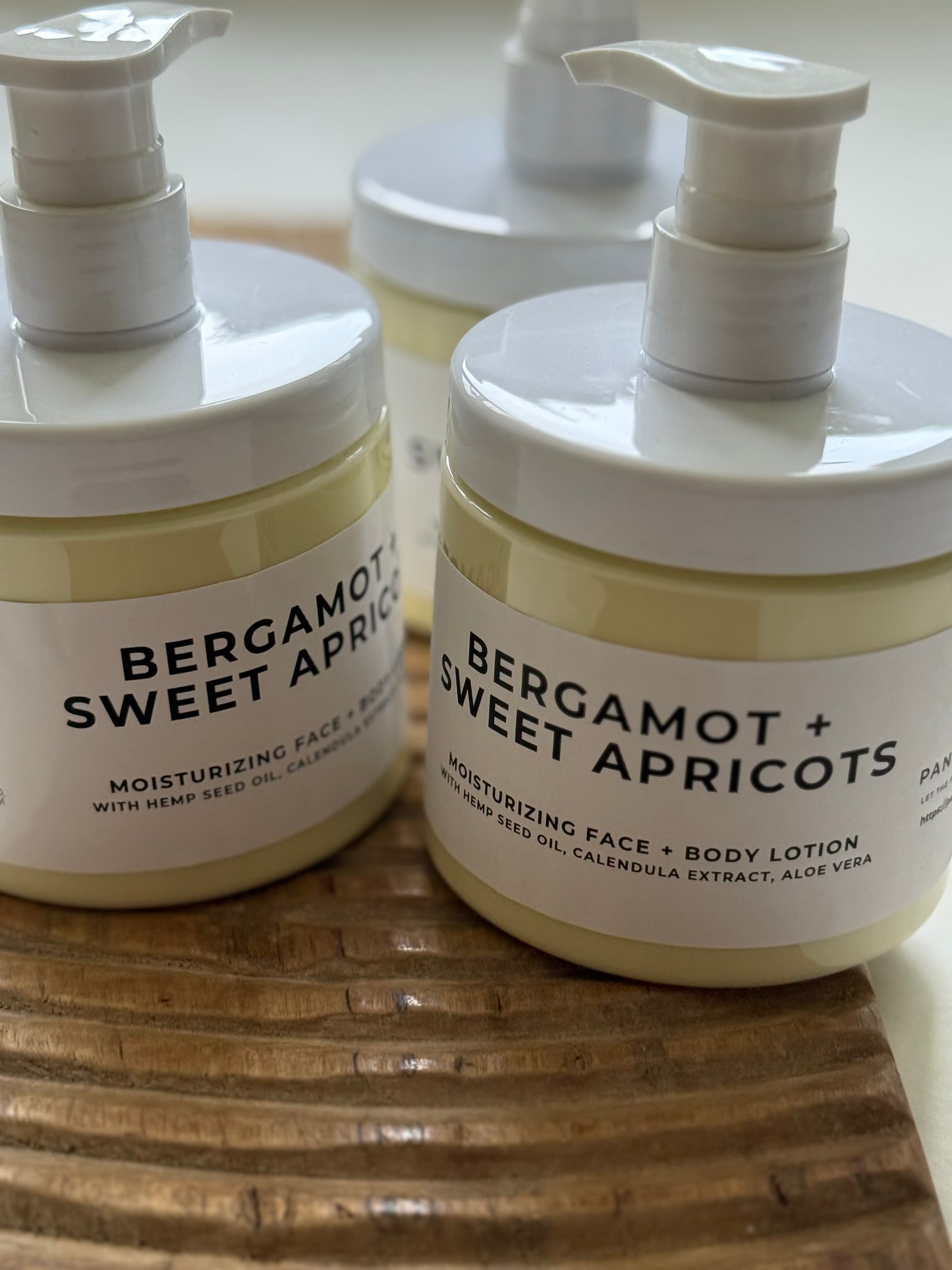 Bergamot + Sweet Apricots moisturizing face and body lotion in pump bottles on a wooden tray.