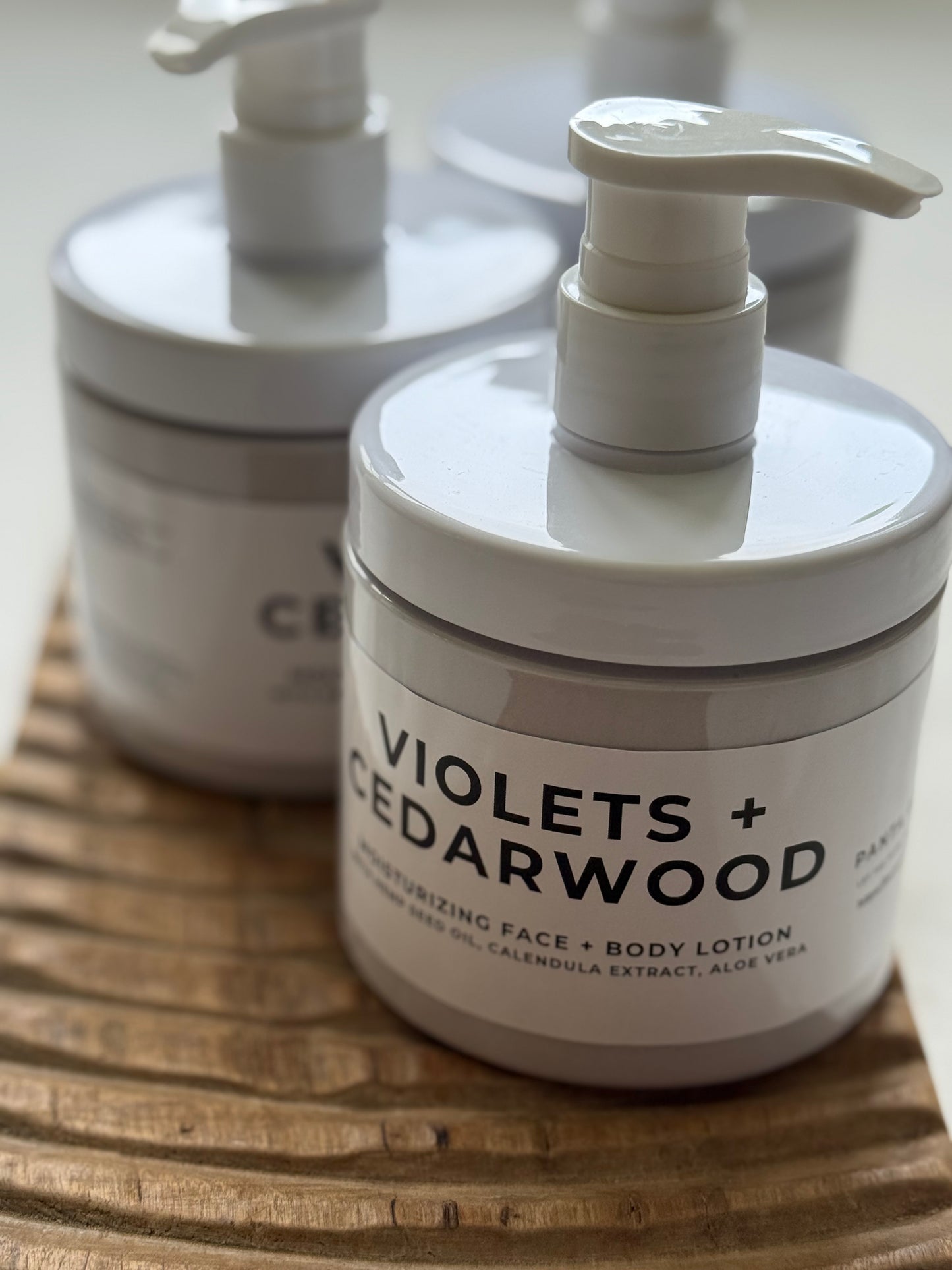 Violets + Cedarwood moisturizing face and body lotion in pump bottles on a wooden tray.