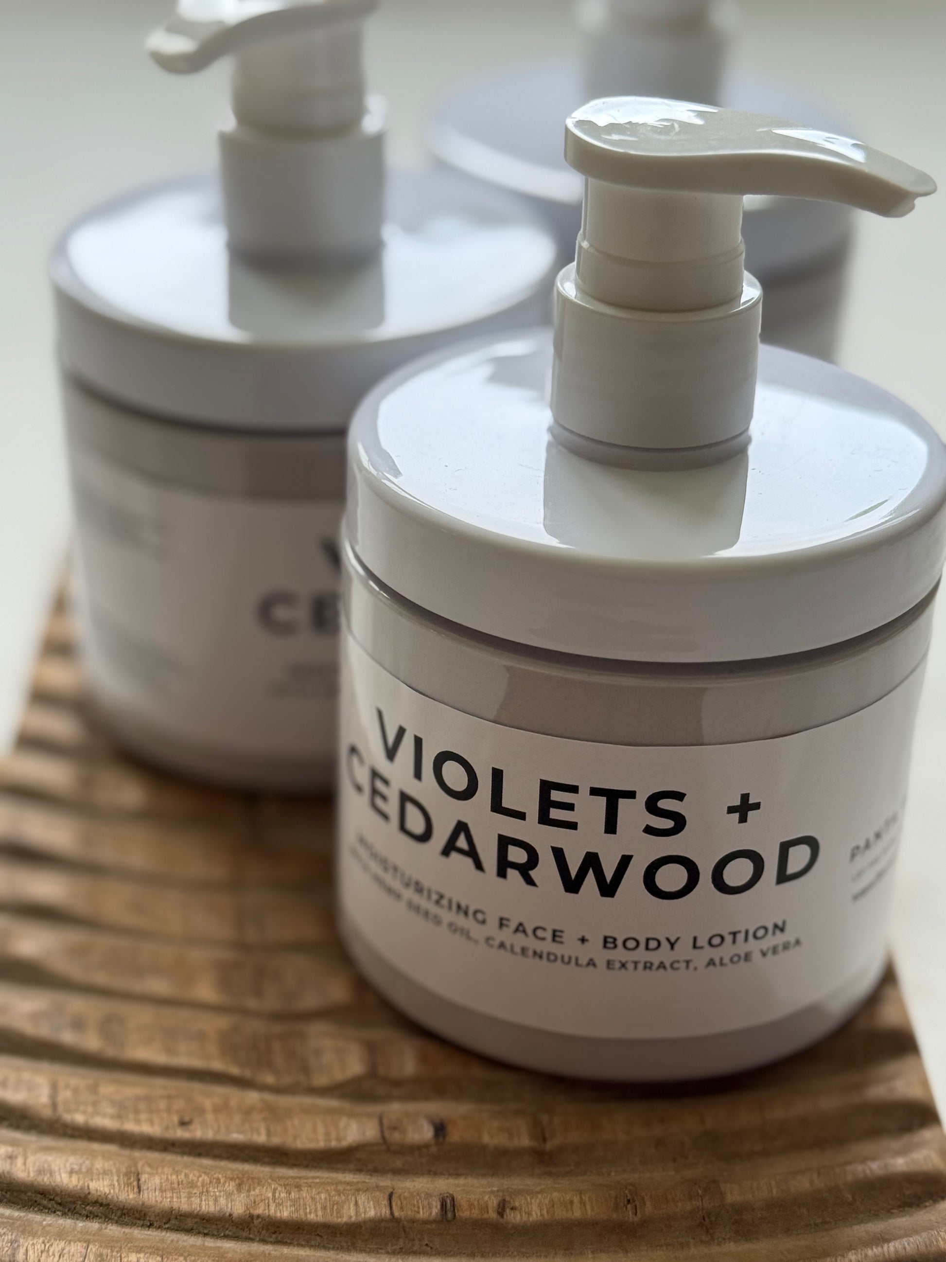 Violets + Cedarwood moisturizing face and body lotion in pump bottles on a wooden tray.