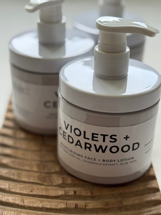 Violets + Cedarwood moisturizing face and body lotion in pump bottles on a wooden tray.