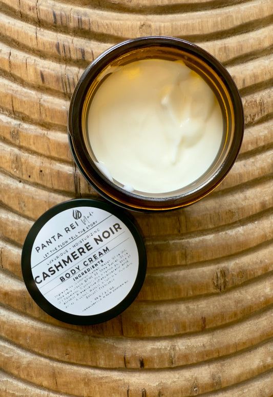 Amber-glass jar of Cashmere Noir Body Cream on carved wooden surface, shown open with creamy texture and matching lid.