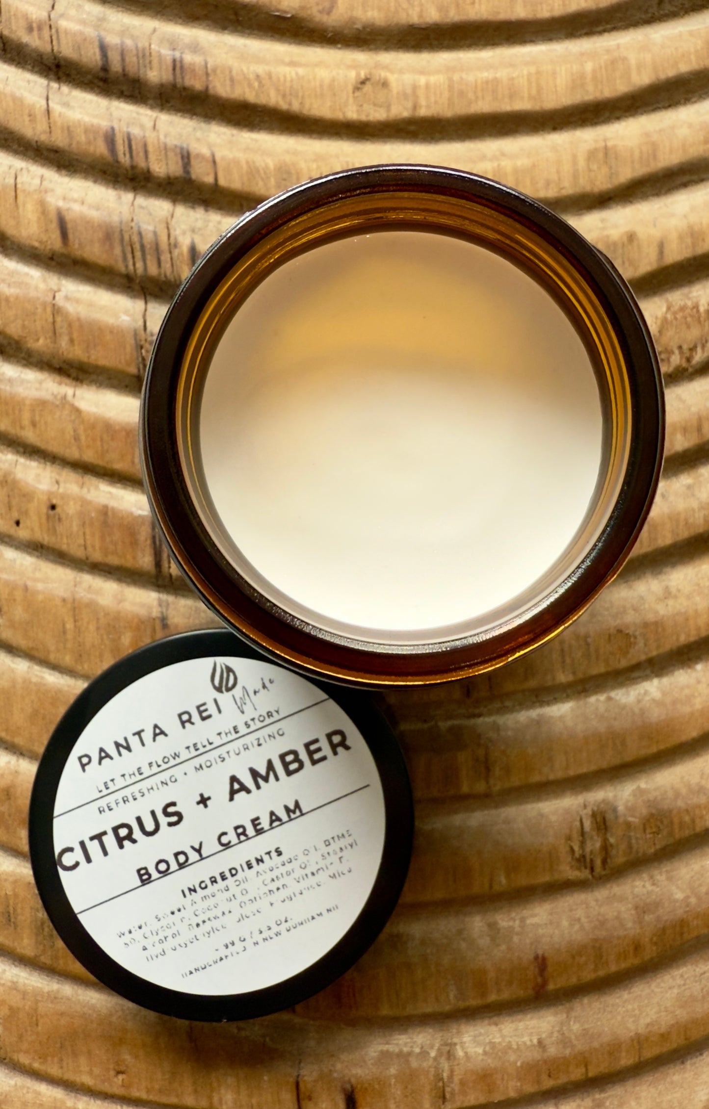 Panta Rei Citrus + Amber body cream jar on a wooden surface. Amber-glass jar of Citrus + Amber Body Cream on carved wooden surface, shown open with soft-peach cream and matching lid.