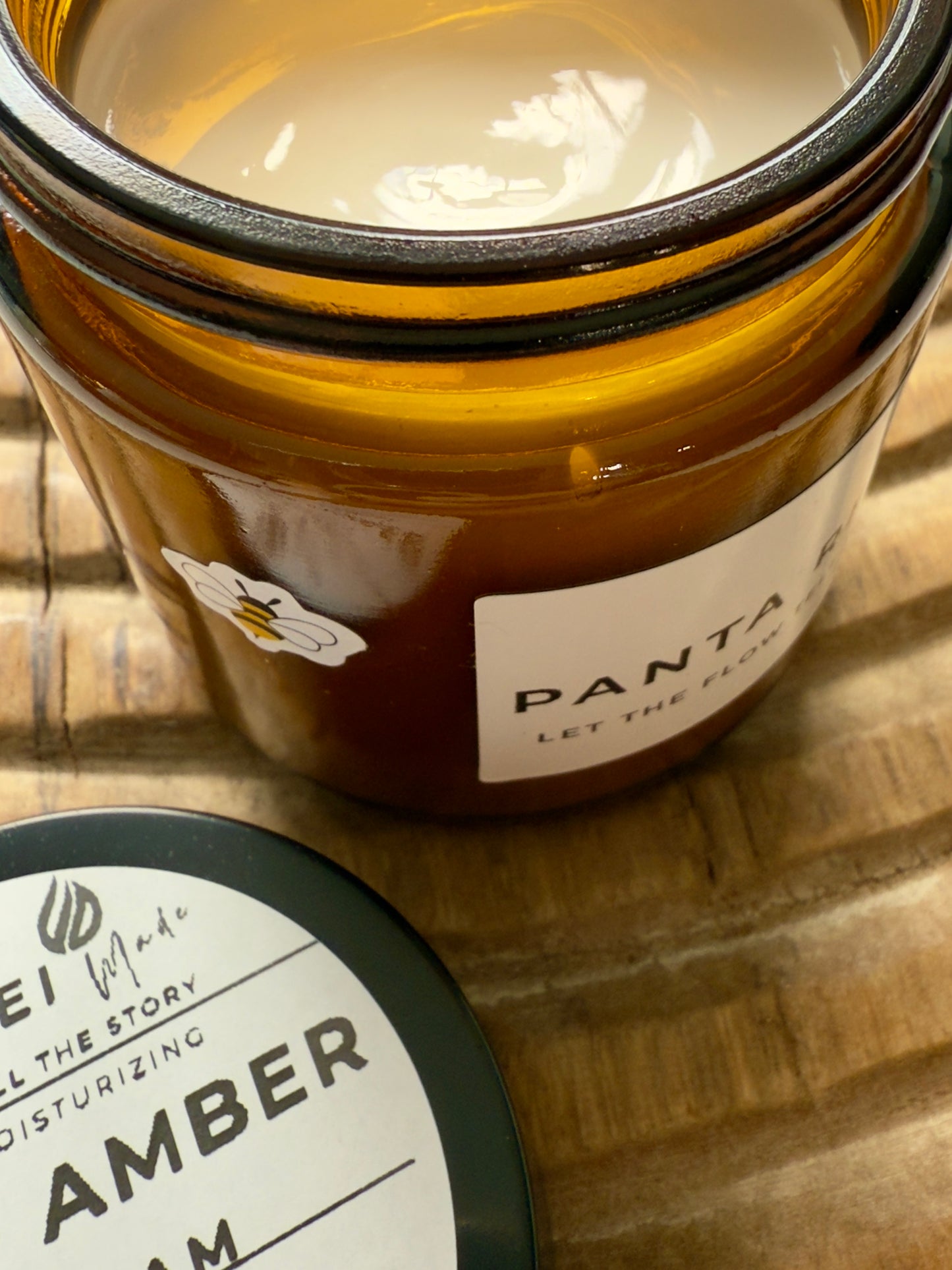 Amber-glass jar of Citrus + Amber Body Cream on carved wooden surface, shown open with soft-peach cream and matching lid.