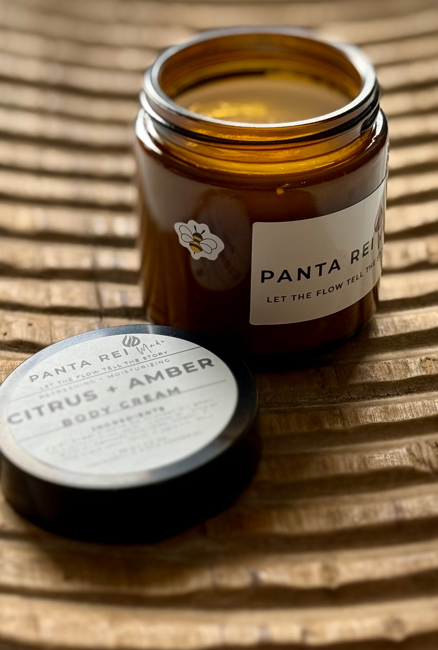Amber-glass jar of Citrus + Amber Body Cream on carved wooden surface, shown open with soft-peach cream and matching lid.