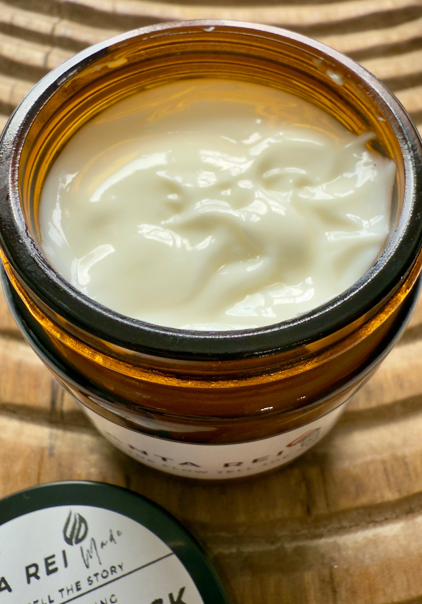 Amber-glass jar of Lavender + Musk Body Cream on carved wooden surface, shown open with smooth cream texture and matching lid.