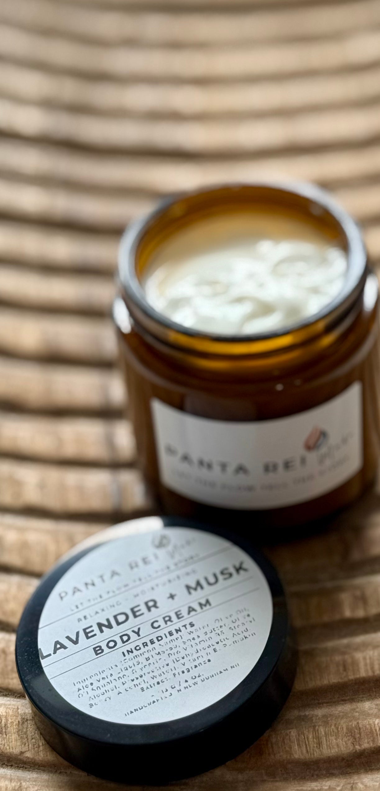 Amber-glass jar of Lavender + Musk Body Cream on carved wooden surface, shown open with smooth cream texture and matching lid.